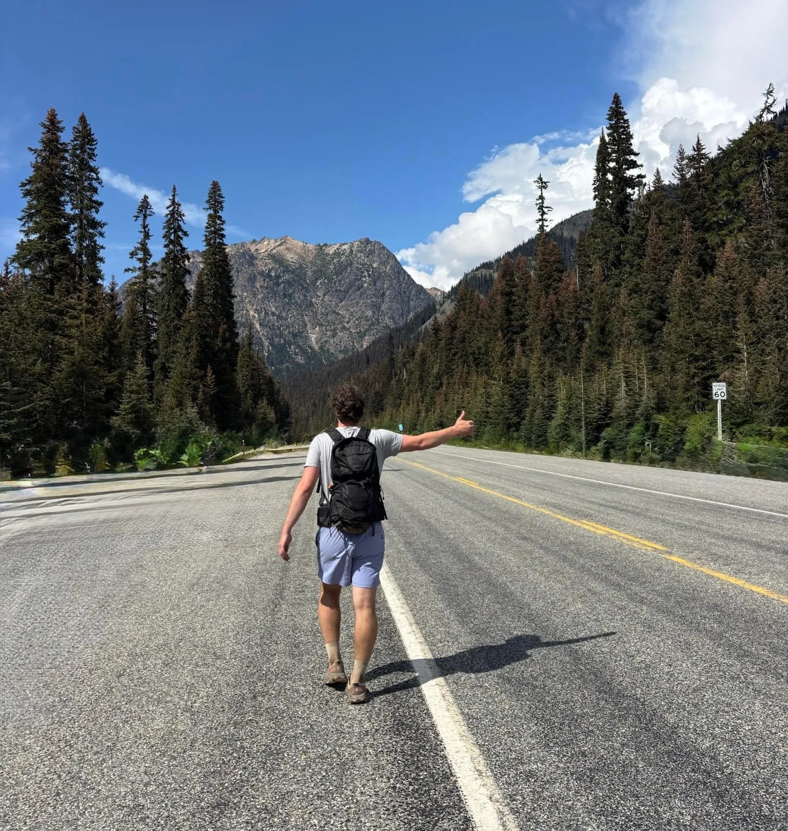 Alden hitchhiking down a mountain highway in the North Cascades with a loaded backpack