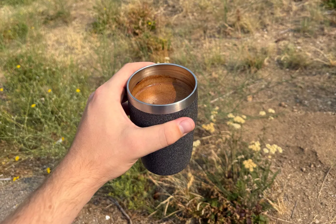 A finished cup of cowboy coffee in an insulated mug, held up over the dry grass and haze in the Boise National Forest